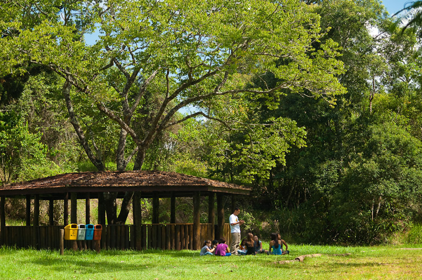Crianças sentadas em roda em um campo recebendo aula de um professor em pé. Ao fundo, um quiosque de madeira