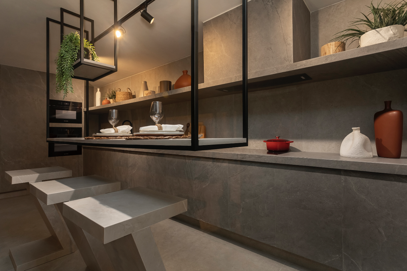 Sala de jantar moderna com bancos retangulares de cimento e mesa suspensa por estrutura de ferro. Ao lado, armário com vasos e plantas